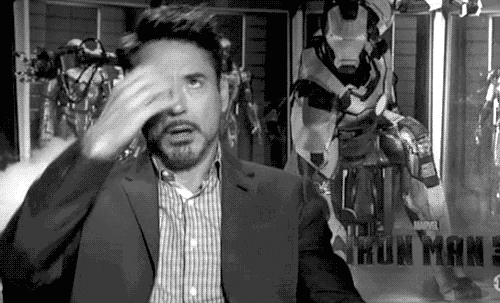GIF em preto e branco de Robert Downey Jr. revirando os olhos e passando a mão na testa em sinal de exaustão, com armaduras do Homem de Ferro ao fundo.