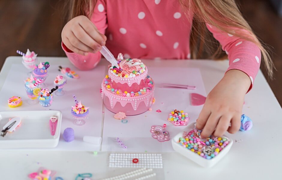 Criança decorando um bolo cor-de-rosa com confeitos coloridos, rodeada de pequenas sobremesas e materiais de artesanato, estimulando a criatividade.