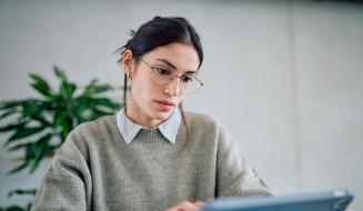 Mulher estudando com tablet em ambiente moderno, usando óculos e blusa cinza, com expressão concentrada e fundo de parede clara e planta verde ao fundo.