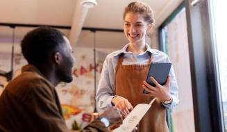 Garçonete sorridente de avental atende cliente em restaurante, segurando tablet enquanto anota o pedido pelo cardápio