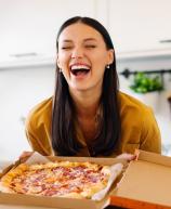 Mulher sorridente segurando uma caixa de pizza em uma cozinha moderna, transmitindo alegria e satisfação ao comer pizza.