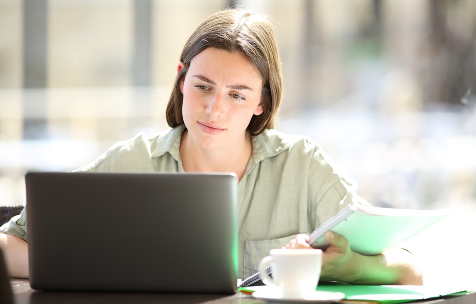 Uma jovem mulher concentrada estudando no computador em um café, com caderno e caneta ao lado, refletindo foco e produtividade. Ambiente iluminado e aconchegante.