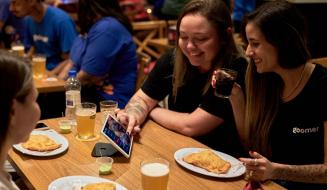 Grupo de amigos em um bar, sorrindo e interagindo com um tablet enquanto desfrutam bebidas e petiscos na mesa, criando momentos de descontração e alegria.