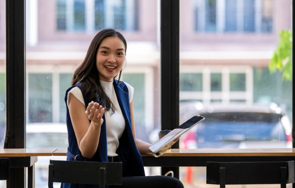 Mulher sorridente em um ambiente de trabalho moderno, sentada em uma mesa com um bloco de notas e um tablet, expressando entusiasmo e confiança.