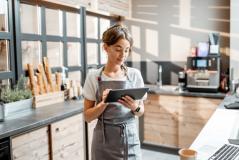 Jovem mulher atendente em cafeteria usando tablet, serviço de atendimento ao cliente, profissionalismo na cozinha com ambiente moderno e aconchegante.