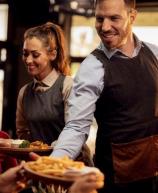 Garçom e garçonete sorrindo servindo pratos de comida e bebida a clientes em um restaurante com ambiente aconchegante.