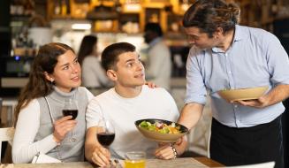 Família apreciando jantar em restaurante, com o garçom atendendo e servi com pratos e taças de vinho, ambiente acolhedor e descontraído.