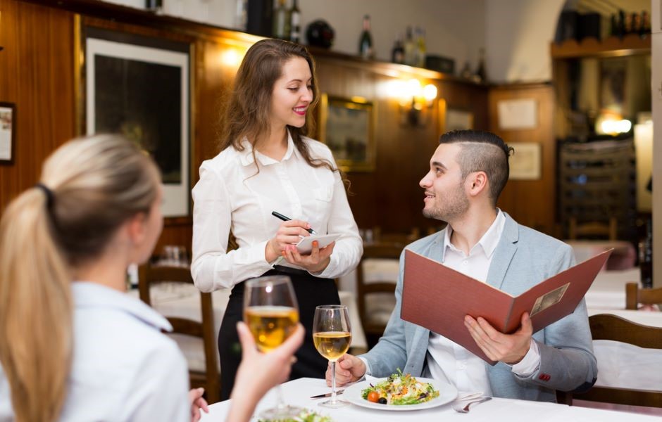 Jovem mulher atendendo clientes em um restaurante elegante, sorrindo e anotando pedidos, enquanto o homem na mesa olha para ela com interesse, com pratos e taças de vinho branco na mesa.
