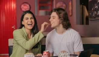 Casal sorridente compartilhando um momento divertido em uma lanchonete, saboreando comida e bebida, com um ambiente descontraído ao fundo.