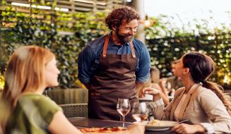 Homem com barba e cabelo cacheado atendendo duas mulheres em um restaurante ao ar livre, com pratos de comida e taças de vinho, ambiente acolhedor e natural.