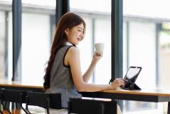 Jovem mulher sorridente utilizando tablet na cafeteria com grande janela de vidro e luz natural, fazendo uma pausa e bebendo chá ou café, promovendo bem-estar e produtividade.