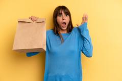 Mulher surpresa segurando sacola de papel com expressão de espanto contra fundo amarelo, representando entrega de comida ou compras online.