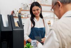 Jovem mulher sorridente fazendo pagamento com smartphone em uma cafeteria, usando uniforme e atendente ao lado, ambiente acolhedor e bem decorado