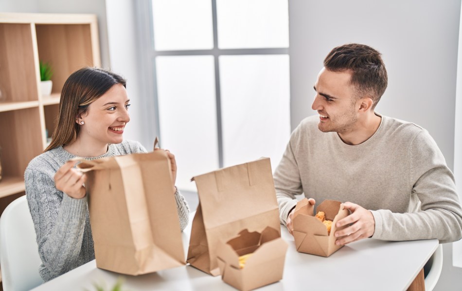 Casal jovem sorridente descompactando embalagens de papel pardo em momento de refeição em casa, ilustrando as tendências de delivery focadas em conveniência e sustentabilidade.