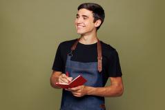 Jovem adulto sorrindo usando avental e caderno, representando empreendedorismo ou trabalho empreendedor em um ambiente interno com fundo neutro.