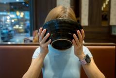 Pessoa segurando um grande prato de comida no rosto em um restaurante, com decoração moderna ao fundo, desfrutando de uma refeição deliciosa.
