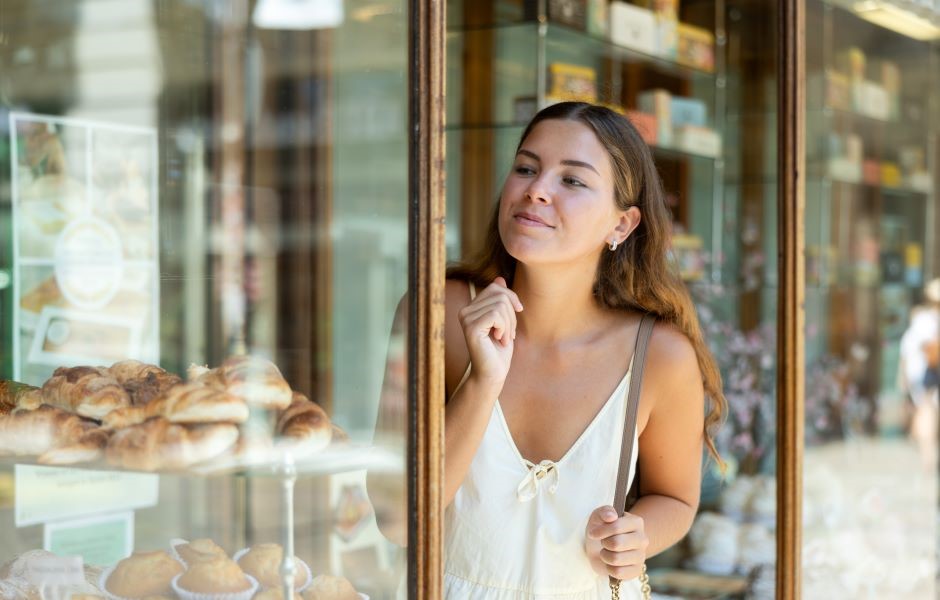 Mulher jovem admirando doces em uma vitrine de padaria ou confeitaria em um dia ensolarado, com expressão de satisfação e curiosidade.