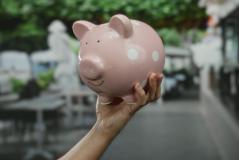 Pessoa segurando uma vaquinha de porcelana rosa com detalhes brancos em um ambiente ao ar livre, simbolizando economia doméstica ou poupança.