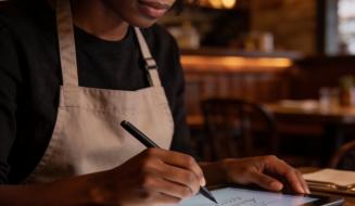 Mulher de avental escreve em tablet com caneta digital, simbolizando fazer a gestão do restaurante a distância do restaurante