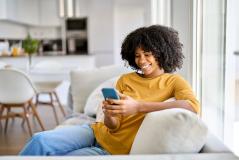 Mulher sorrindo usando celular no sofá em casa, ambiente moderno e aconchegante com iluminação natural, ideal para postar sobre comunicação e tecnologia.
