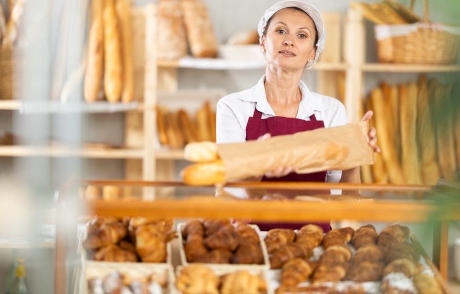 A profissional de panificação apresenta um pão fresco em uma padaria com uma variedade de outros produtos de confeitaria ao fundo, ela segura uma sacola biodegradável.