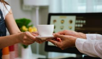 A mulher recebendo uma xícara de café de um atendente em um ambiente interno, com computador ao fundo, simbolizando serviço de cafeteria ou entrega de bebidas