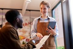 Garçonete sorridente de avental atende cliente em restaurante, segurando tablet enquanto anota o pedido pelo cardápio