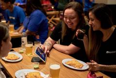 Grupo de amigos em um bar, sorrindo e interagindo com um tablet enquanto desfrutam bebidas e petiscos na mesa, criando momentos de descontração e alegria.