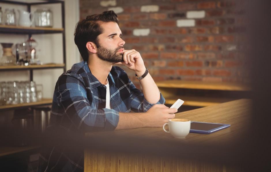 Homem jovem de barba e camisa xadrez azul, sentado em uma mesa de café. Ele apoia a mão no queixo e olha para o lado com expressão séria. À sua frente estão um celular na mão, uma xícara de café e um tablet.