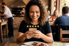 Cliente com cabelos cacheados sorri ao usar o celular para avaliar com cinco estrelas sua experiência no restaurante. Sentada à mesa com um prato de churrasco e uma bebida, ela aproveita o ambiente movimentado ao fundo, onde funcionários atendem outros clientes próximos à churrasqueira.