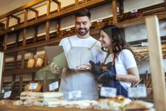 Duas pessoas no balcão de uma padaria, um homem e uma mulher, sorrindo e observando uma tabela de produtos, com prateleiras de pães ao fundo, demonstrando uma conversa descontraída.