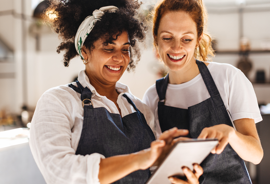 Duas mulheres sorrindo e olhando para um tablet em uma cozinha moderna, vestindo aventais pretos, o que indica uma aula de culinária ou cozinha colaborativa.