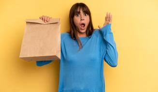 Mulher surpresa segurando sacola de papel com expressão de espanto contra fundo amarelo, representando entrega de comida ou compras online.