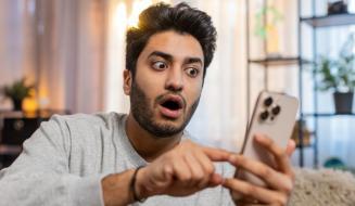 Homem surpreso usando celular em casa, ilustrando a experiência de diversão e surpresa ao usar um smartphone com tecnologia moderna.