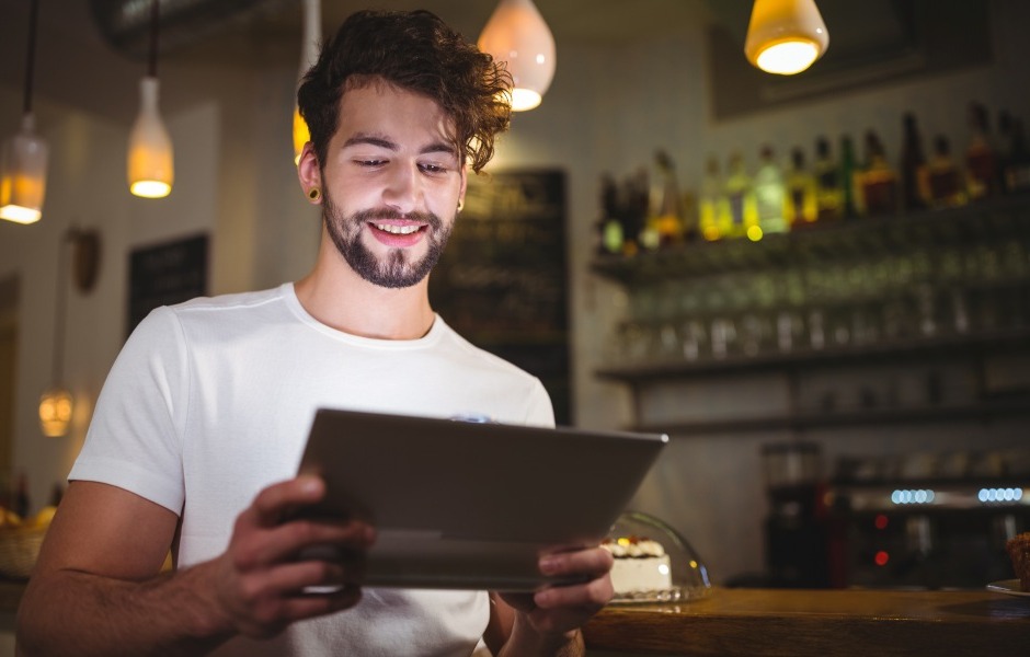 Homem sorridente em uma cafeteria ou bar, segurando e olhando para um cardápio digital em um tablet.