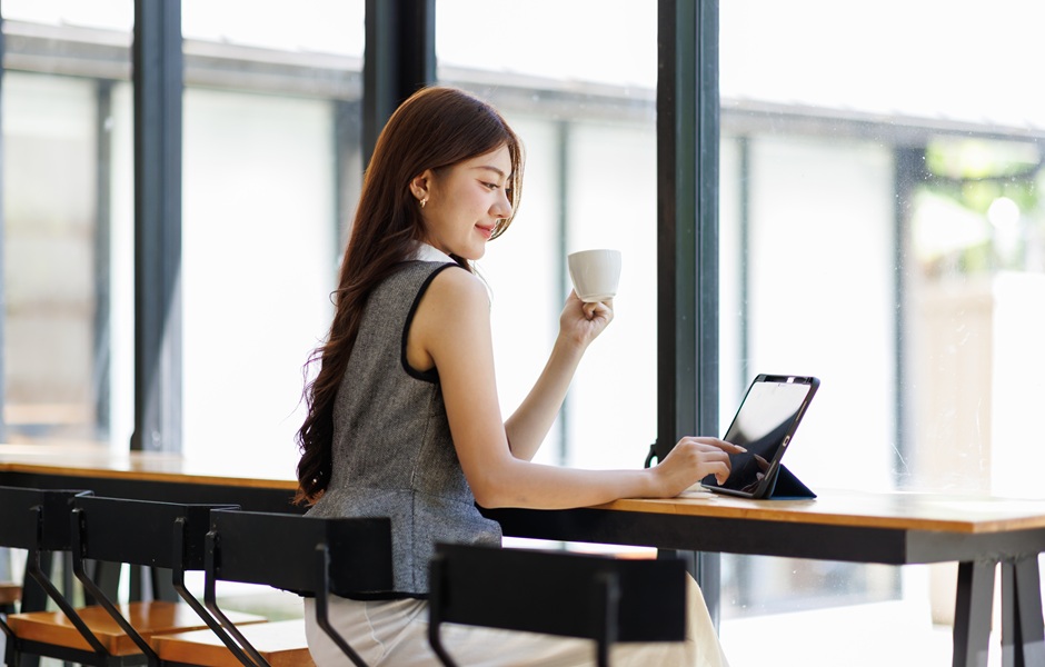 Jovem mulher sorridente utilizando tablet na cafeteria com grande janela de vidro e luz natural, fazendo uma pausa e bebendo chá ou café, promovendo bem-estar e produtividade.