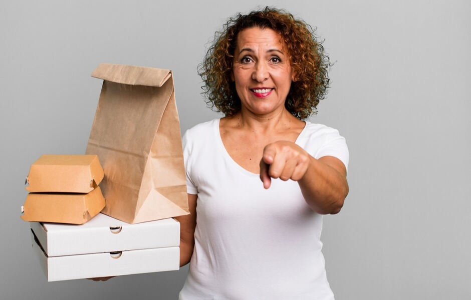 Mulher sorridente segurando embalagens de comida e apontando para a câmera, transmitindo uma sensação de serviço de entrega. Ideal para destacar opções de delivery.