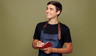 Jovem adulto sorrindo usando avental e caderno, representando empreendedorismo ou trabalho empreendedor em um ambiente interno com fundo neutro.