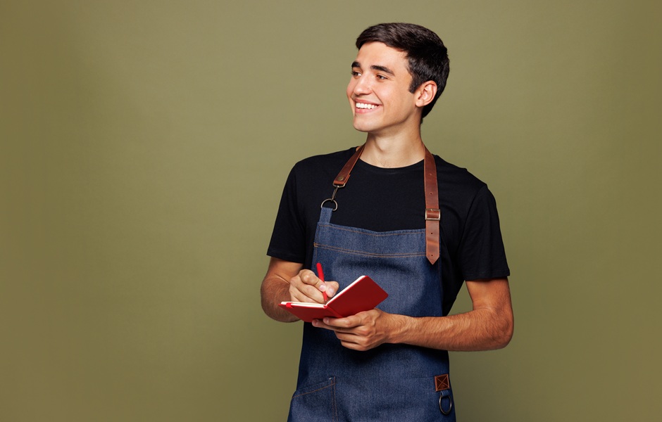 Jovem adulto sorrindo usando avental e caderno, representando empreendedorismo ou trabalho empreendedor em um ambiente interno com fundo neutro.
