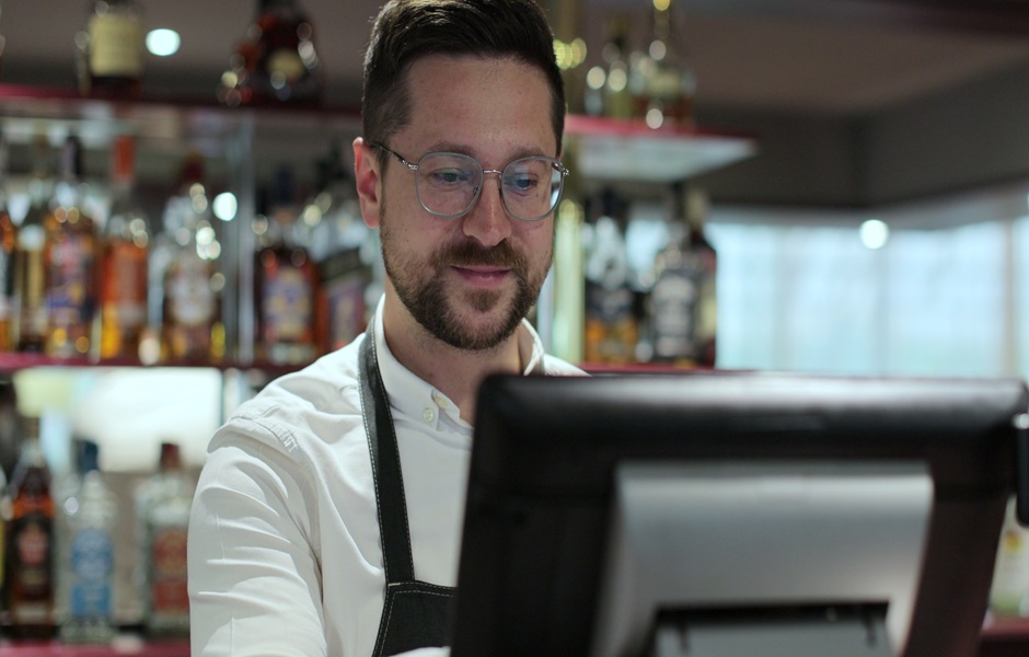 Barista profissional atendendo clientes em uma cafeteria, preparado para fazer pedido, com prateleiras de bebidas alcoólicas ao fundo e ambiente moderno.