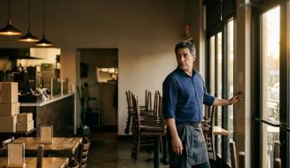 Homem com camisa azul e avental escuro junto à porta de vidro de um restaurante, olhando para trás com expressão pensativa enquanto toca no interruptor de luz. Ao fundo, veem-se cadeiras empilhadas sobre as mesas, caixas de papelão empilhadas e luminárias pendentes acesas. A iluminação natural vinda da janela cria uma atmosfera de encerramento de expediente no estabelecimento.