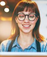 Mulher sorridente com óculos de armações pretas e cabelo castanho, usando camisa xadrez azul, em ambiente de cafeteria, transmitindo alegria e satisfação.
