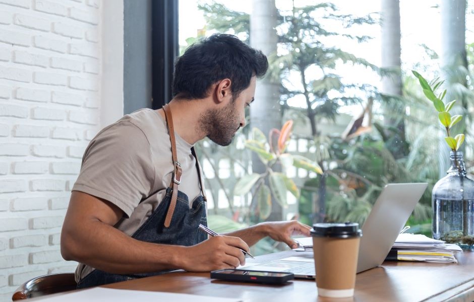 Homem estudando e fazendo anotações com laptop, celular, caneta e café na mesa de um espaço moderno, com vista para a natureza através de uma grande janela.