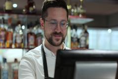 Barista profissional atendendo clientes em uma cafeteria, preparado para fazer pedido, com prateleiras de bebidas alcoólicas ao fundo e ambiente moderno.