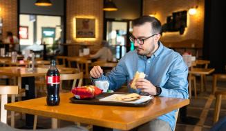 Homem com barba e óculos tomando café da manhã com pão e refrigerante em cafeteria moderna e aconchegante.