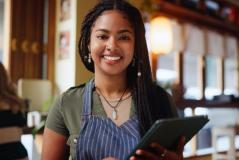 Mulher sorridente com cabelo trançado, usando avental no ambiente de um restaurante ou café, segurando um tablet, no contexto de trabalho na área de gastronomia ou atendimento ao cliente.
