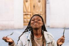 Mulher sorridente segurando garfo e faca ao ar livre, desfrutando uma refeição, com expressão de felicidade, em ambiente urbano. Uma cena que transmite alegria e satisfação ao comer.