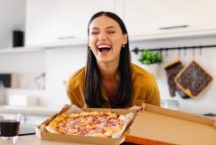 Mulher sorridente segurando uma caixa de pizza em uma cozinha moderna, transmitindo alegria e satisfação ao comer pizza.