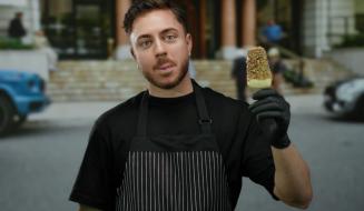 Jovem chef com barba e cabelo curto, vestindo avental preto listrado e luvas pretas, segurando um picolé de chocolate e limão em frente a um restaurante ou café ao ar livre.