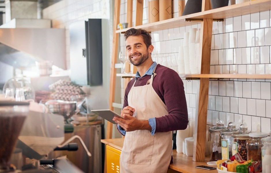 Homem sorridente com barba, vestindo avental e camisa azul, trabalhando em uma cafeteria ou café, segurando um tablet com prateleiras de produtos ao fundo.